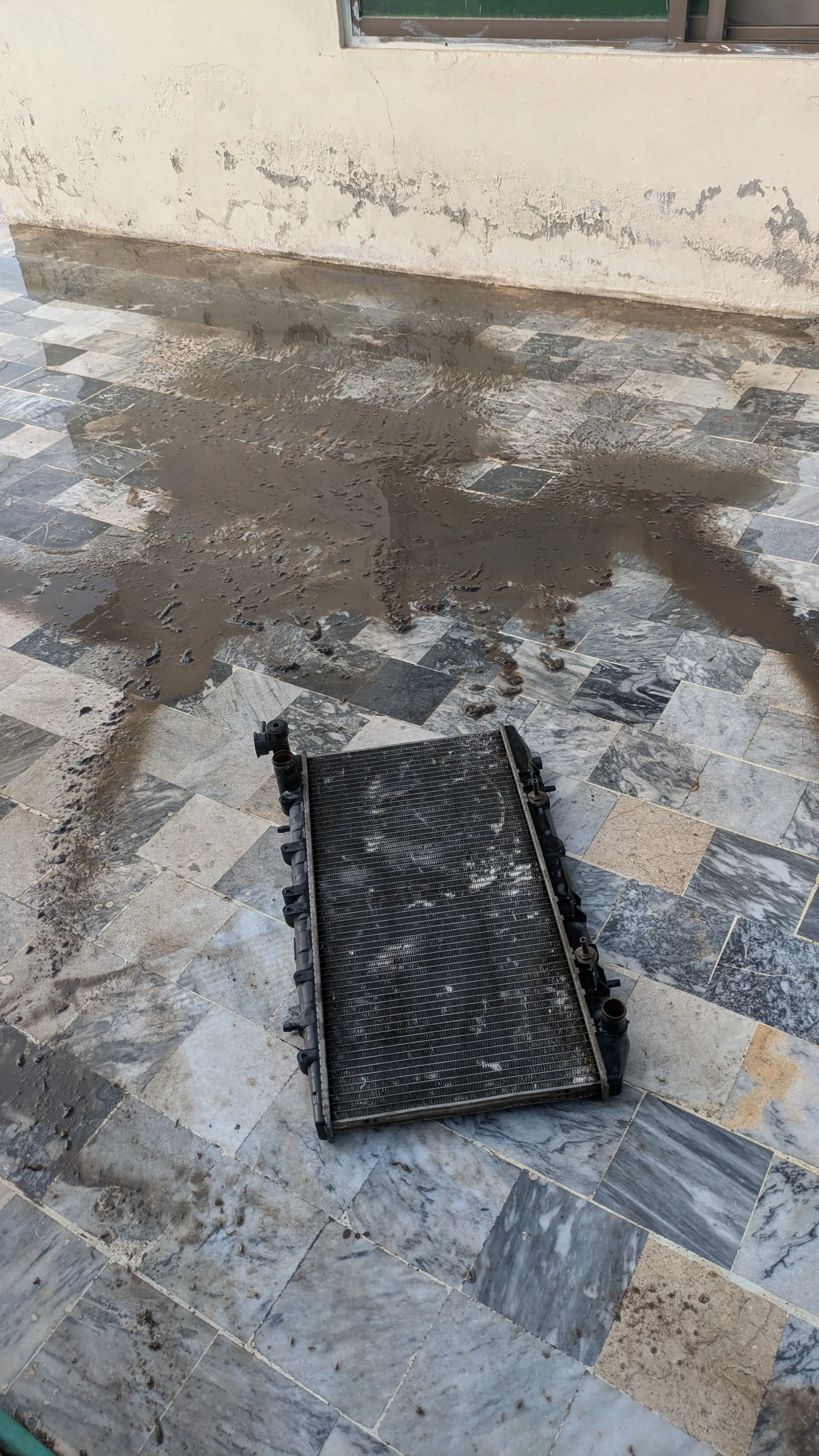 The Nissan Sunny's radiator lying on wet tiles after pressure washing, surrounded by a wide spread of brown mud washed out of its fins.